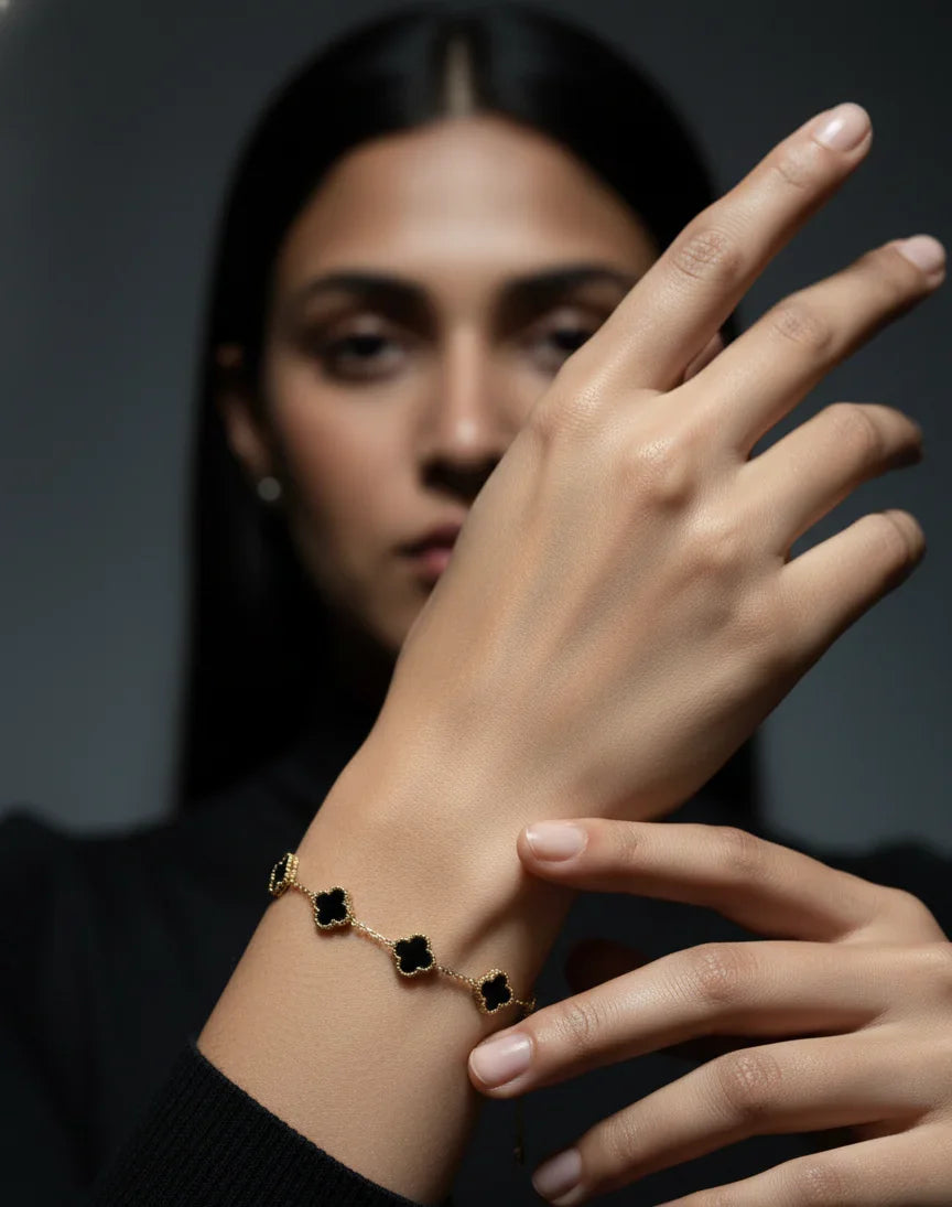 Woman wearing a gold bracelet with black clover charms, hand raised, on dark background